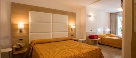 Quarto de hotel com uma cama grande, uma cama menor, uma escrivaninha com abajur e uma cadeira.