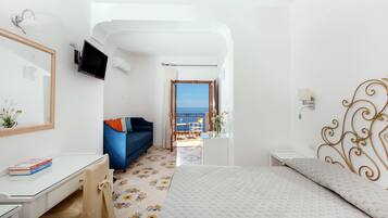 A hotel room with a bed, a desk, a chair, a TV mounted on the wall, and a view of the sea through an open door.