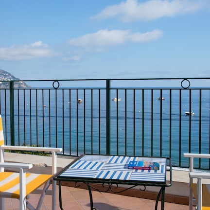 A terrace with a table and chairs overlooking the sea, featuring a view of boats and a mountainous coastline.
