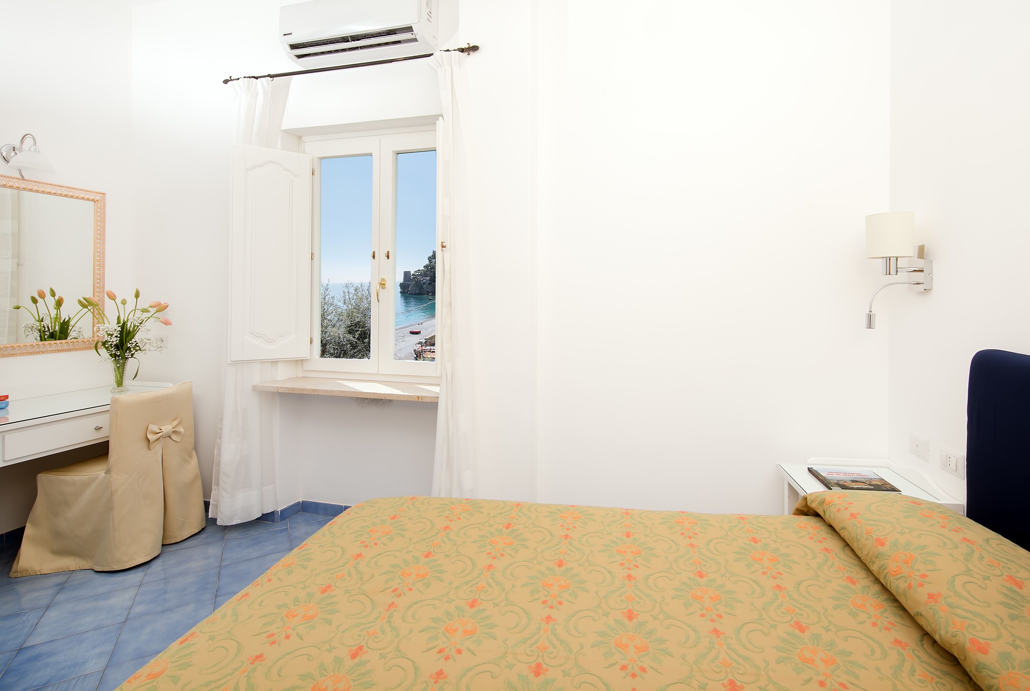 A bedroom with a bed, a window with a view, a bedside table, a mirror, and a wall-mounted lamp.