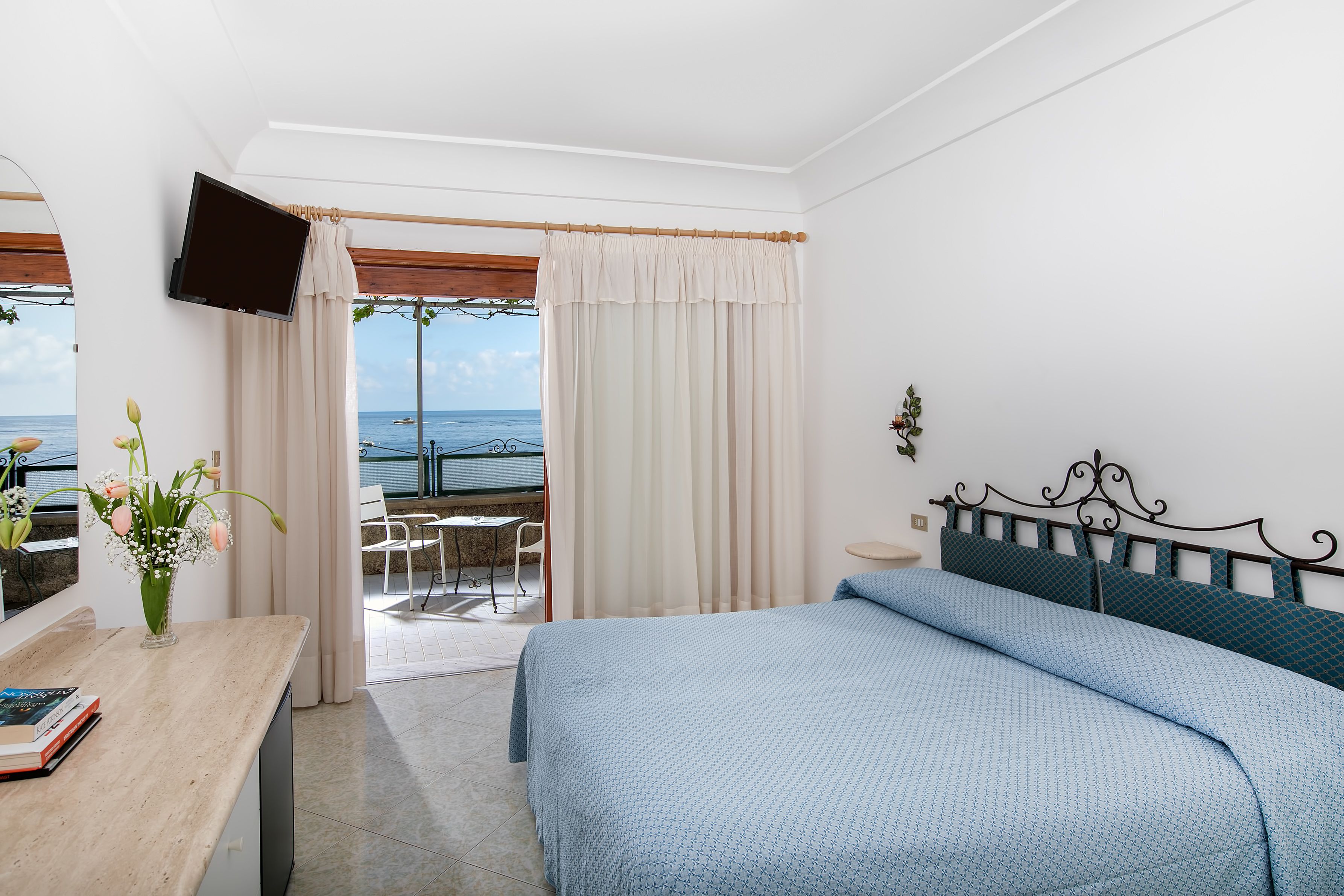 A bedroom with a bed, a TV, a desk with a vase of flowers, and a view of the sea through a window.