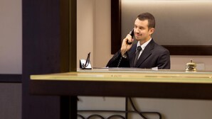 A man in a suit speaking into a phone at a reception desk with a nameplate and a telephone. - Hotel dei Mille (Rome)