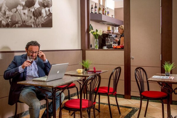 Un hombre sentado a una mesa con una computadora portátil, un jarrón con flores y una máquina de café al fondo.
