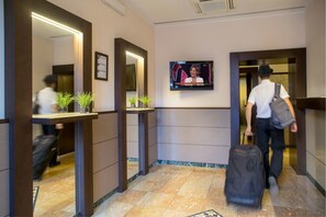 A hotel lobby with a reception desk, a television mounted on the wall, and a person with a suitcase walking towards the elevator. - Hotel dei Mille (Rome)