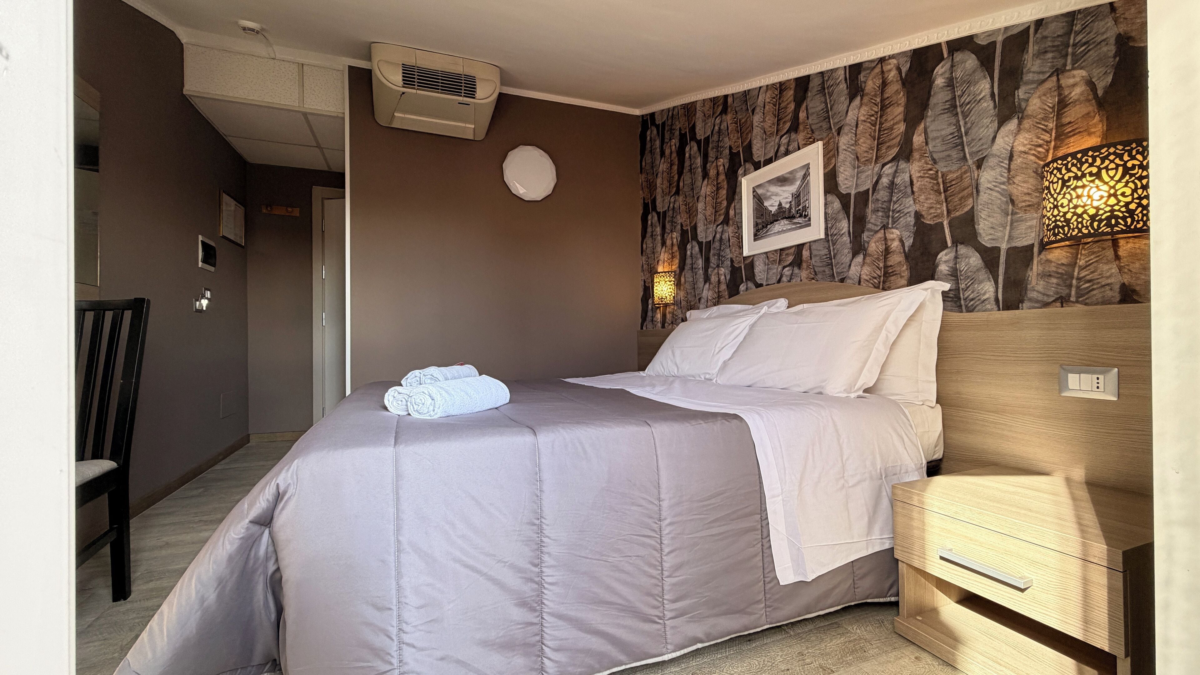 A bedroom with a bed, a nightstand, a wall-mounted air conditioner, a wall-mounted lamp, and a framed picture.