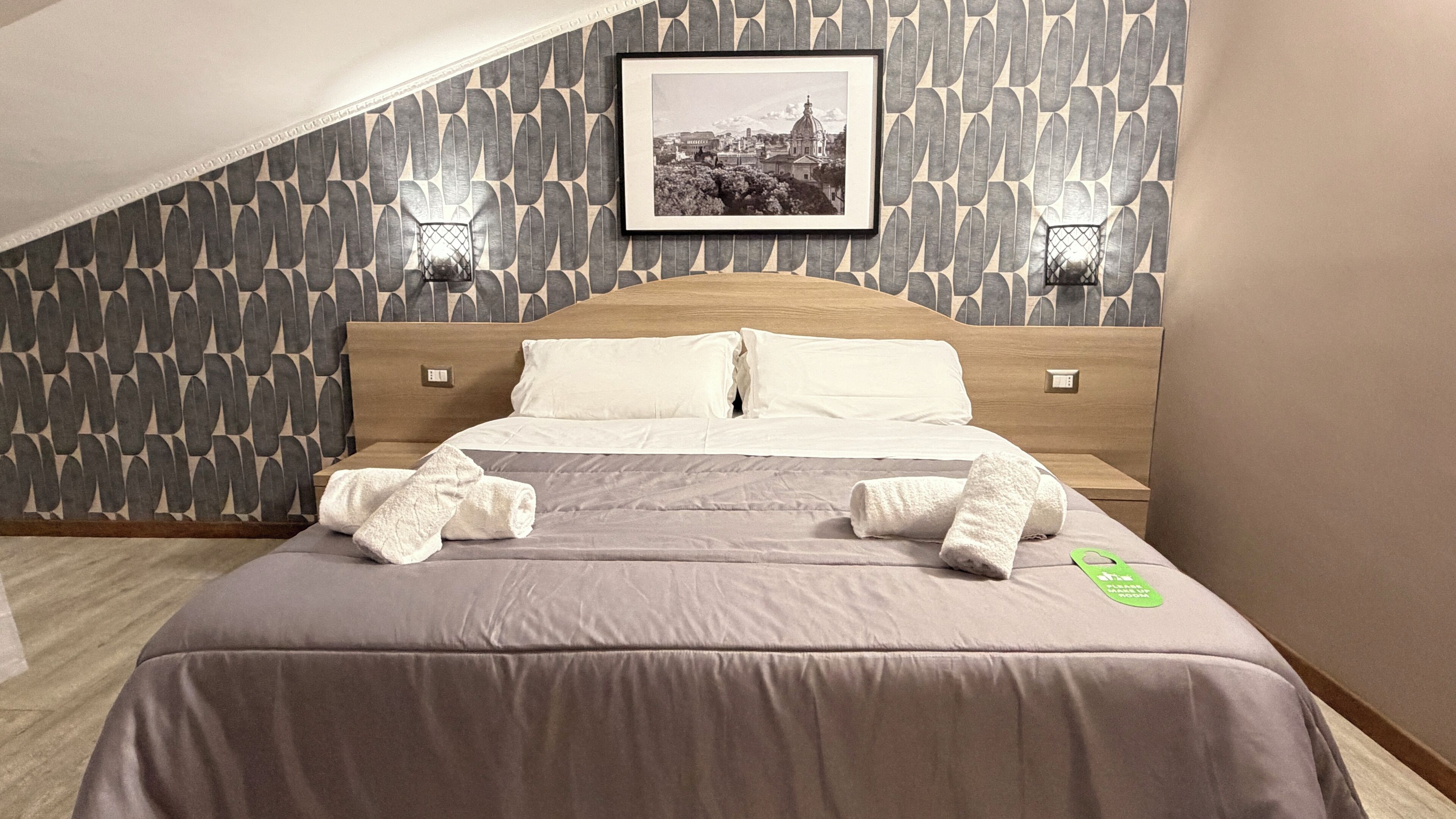 A neatly made bed with white linens and towels, a framed picture on the wall, and a patterned wallpaper.