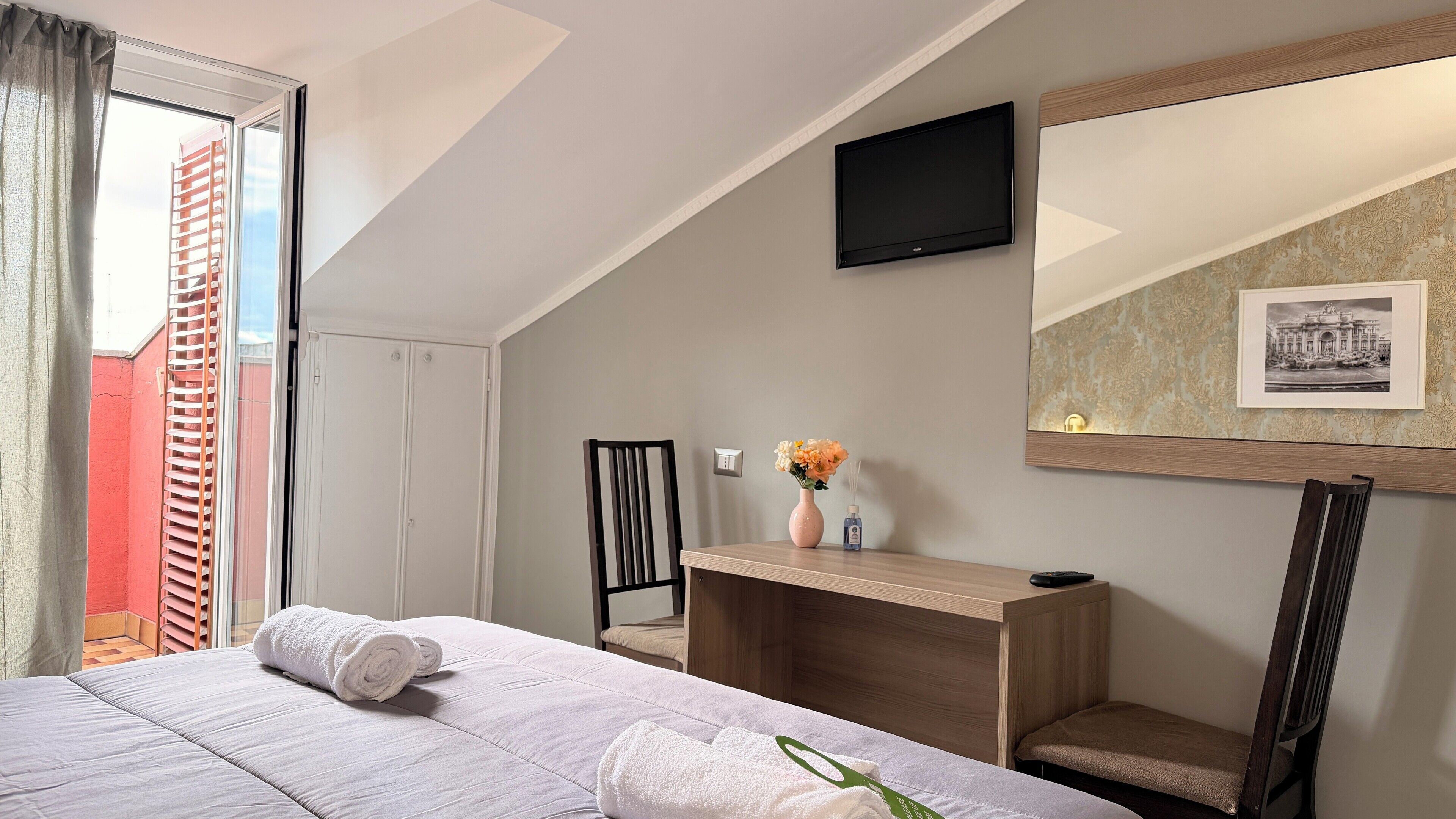 Habitación de hotel con cama, escritorio, silla, televisor montado en la pared y ventana con vista a un edificio rojo.