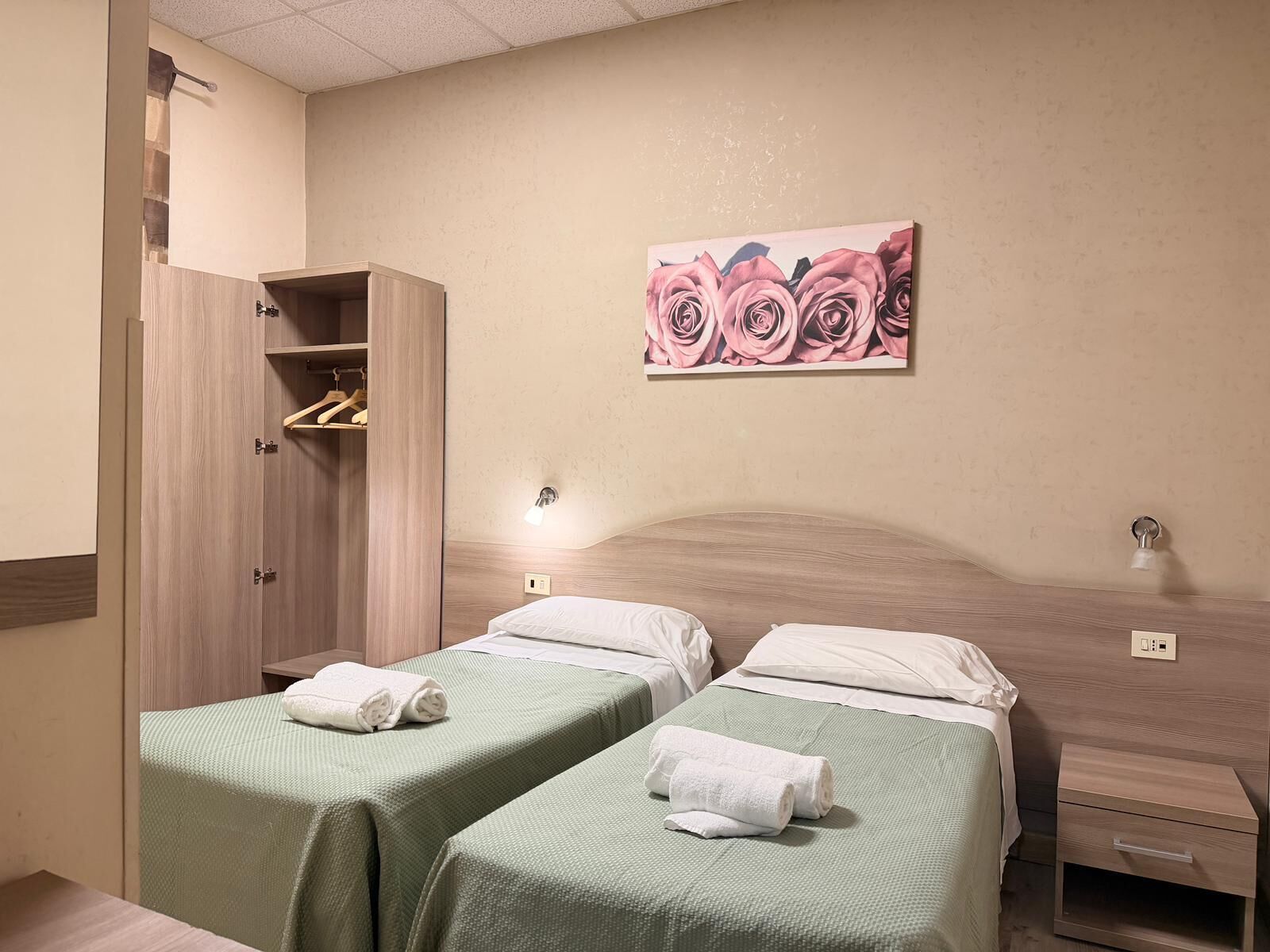 A hotel room with two single beds, a wooden wardrobe, a painting of roses, and a bedside table with a lamp.