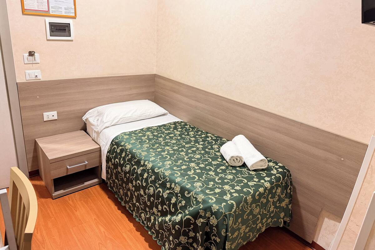 A small, neatly made hotel room with a single bed, a nightstand, a chair, and a wall-mounted electrical outlet.