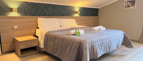 A neatly made bed with a grey bedspread and two rolled towels, a wooden nightstand with a drawer, and a wall-mounted light fixture.