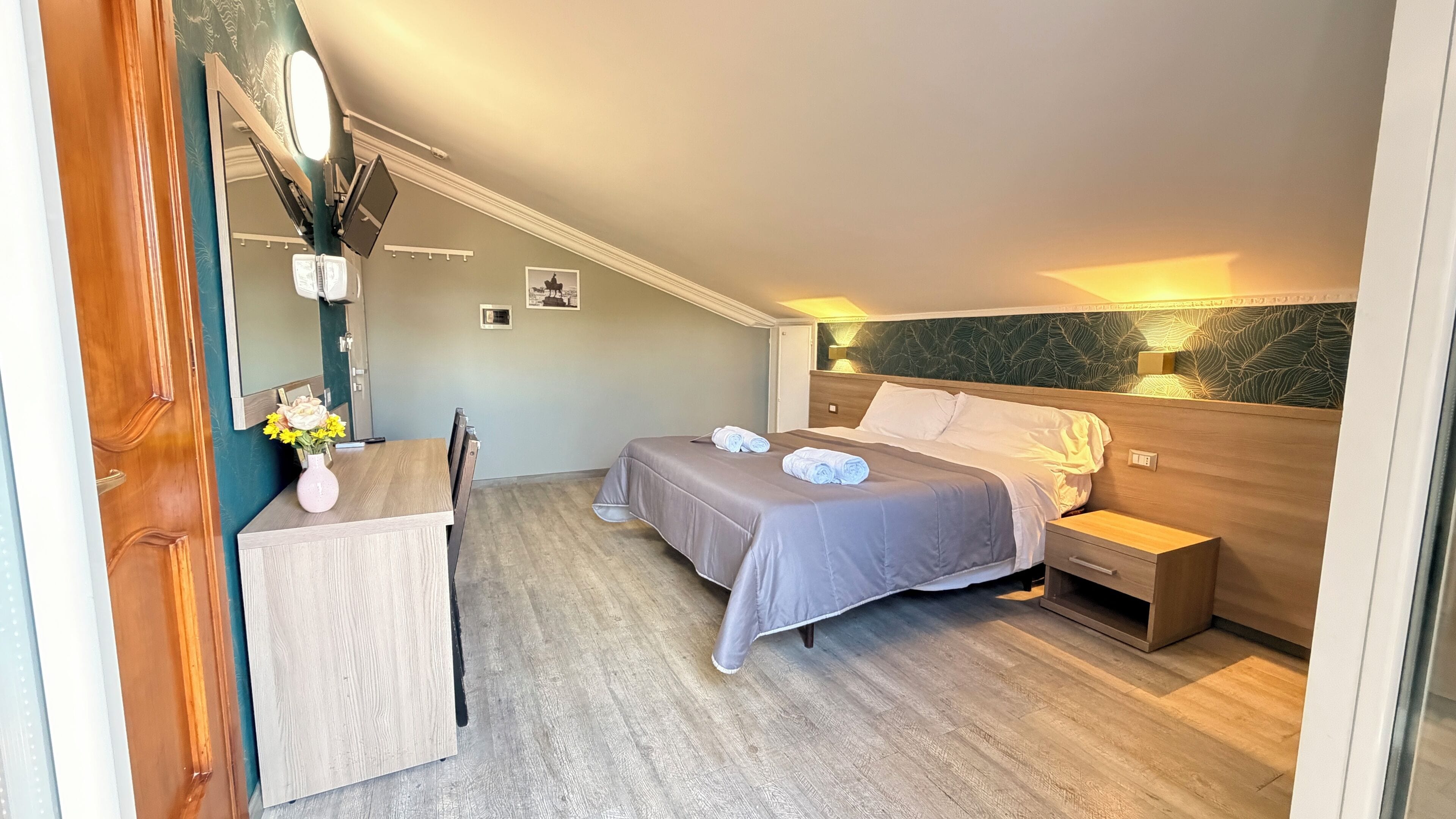A hotel room with a bed, bedside table, chair, desk, and a small table with a vase of flowers.