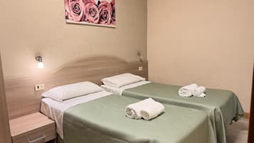 A hotel room with two beds, a nightstand, a wall-mounted light, and a painting of roses.