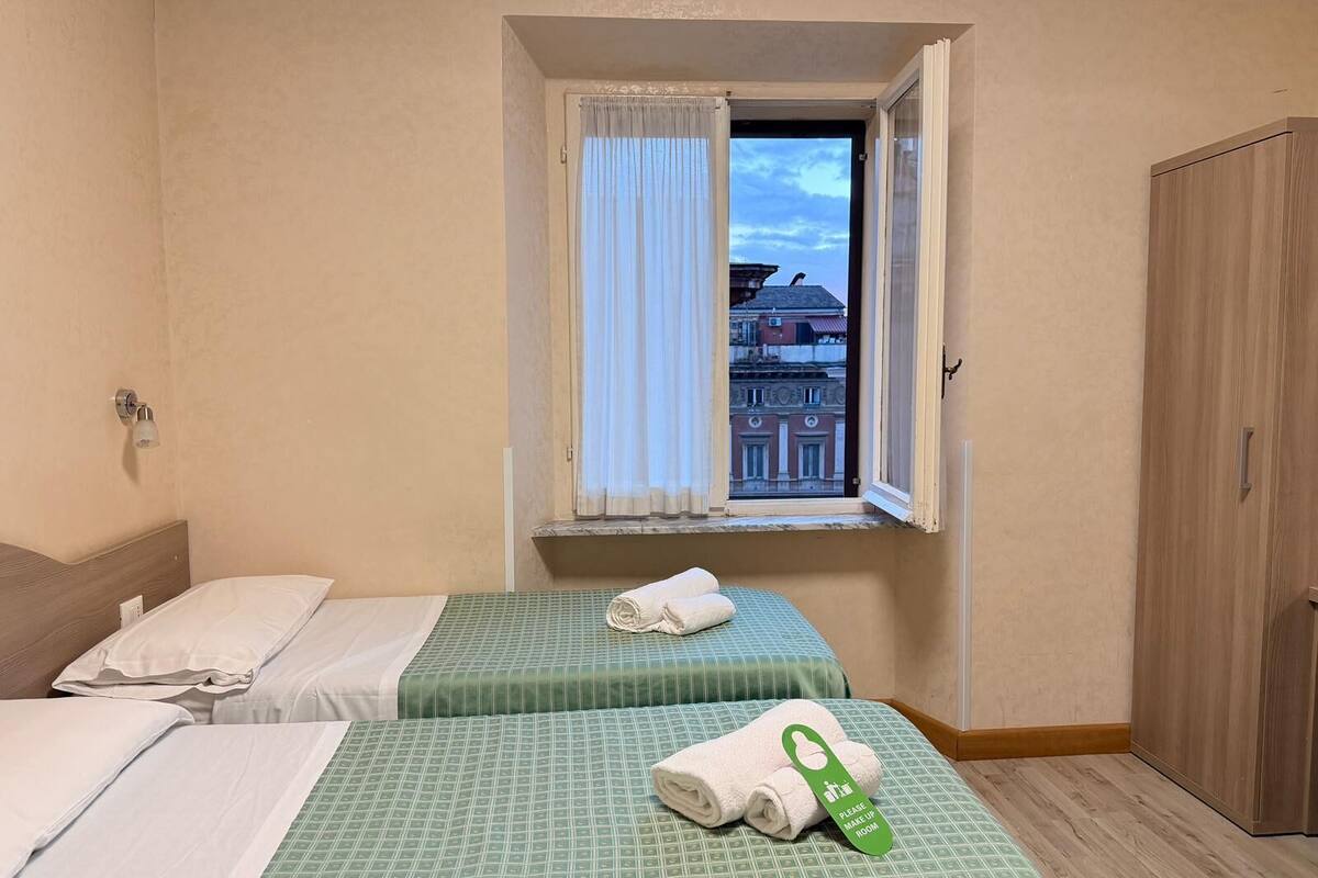 A hotel room with two beds, a window with curtains, a view of buildings, and a wooden wardrobe.