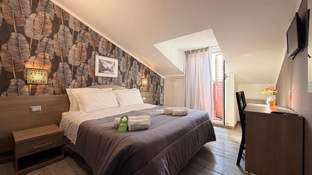 A bedroom with a large bed, a desk, a chair, a nightstand, and a wall-mounted TV.