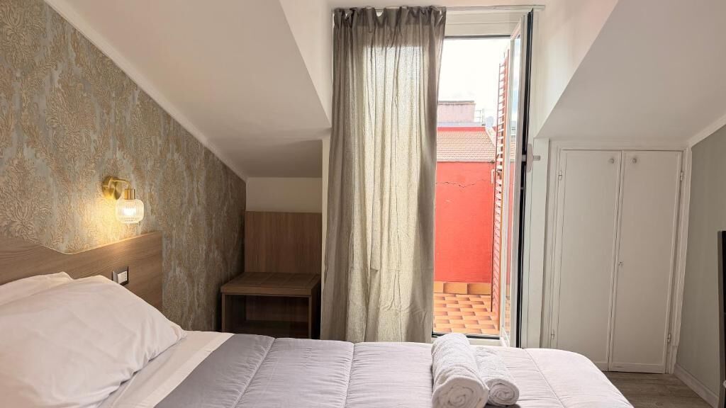 Una camera d'albergo con un letto, un comodino, una lampada a parete, una finestra con tende e vista su un edificio rosso.