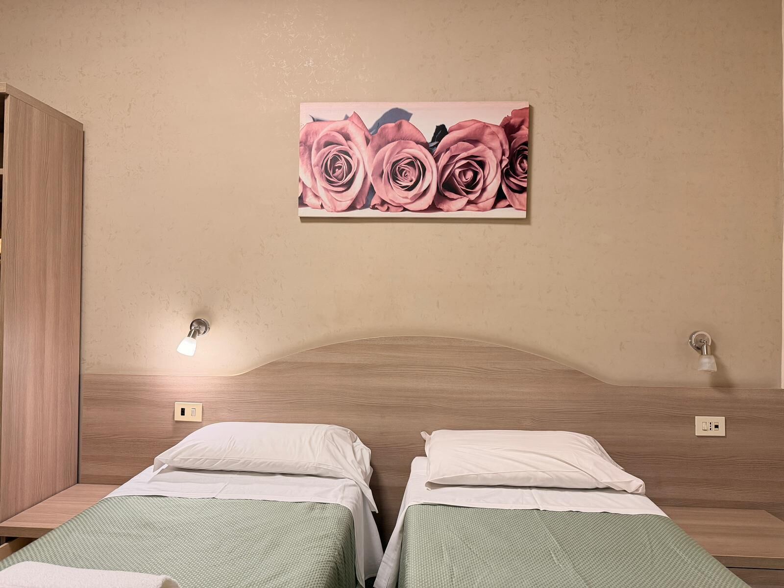 A hotel room with two beds, a wooden headboard, a painting of pink roses, and bedside lamps.
