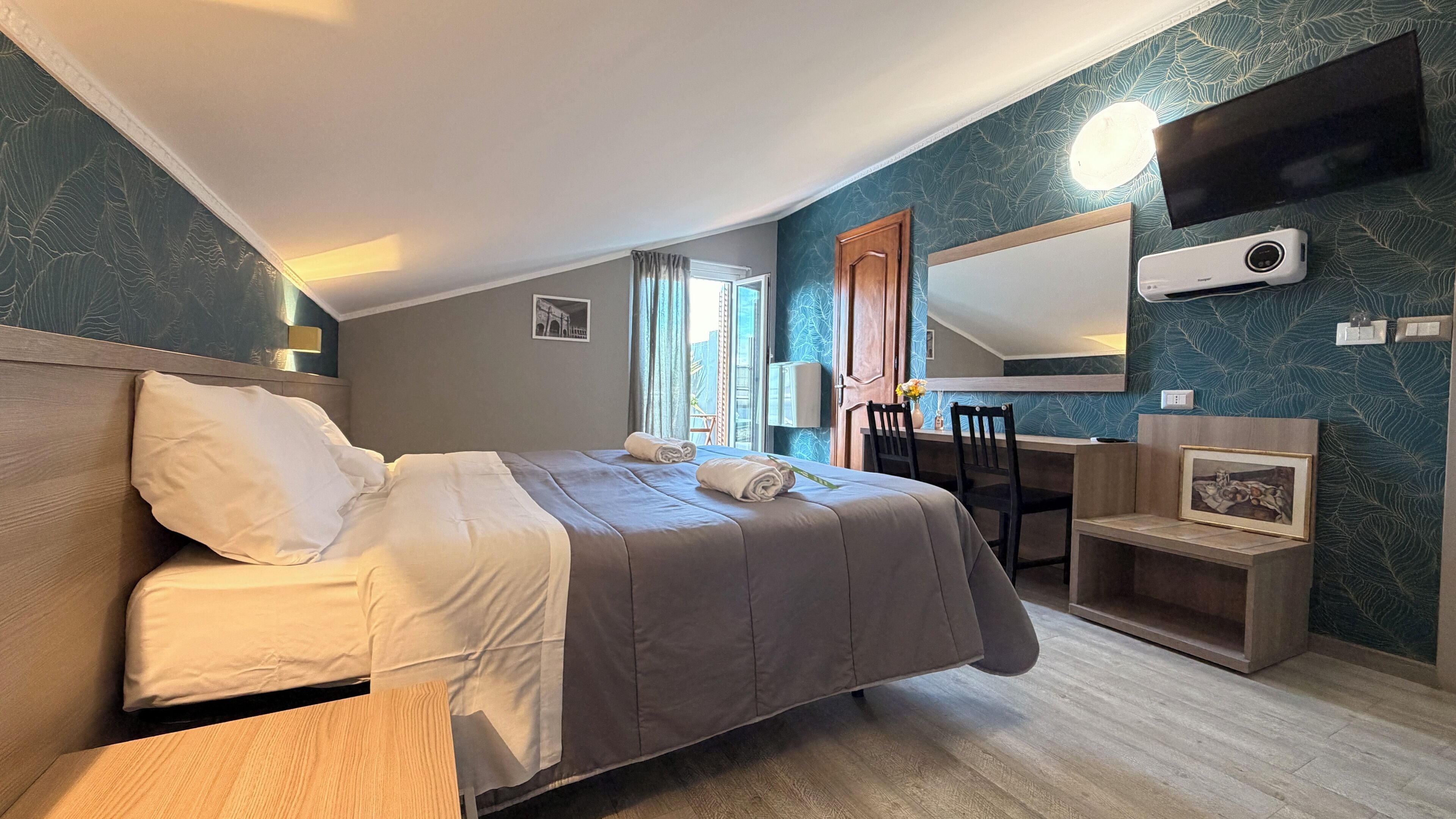 A hotel room with a bed, a desk, a chair, a mirror, and a TV.