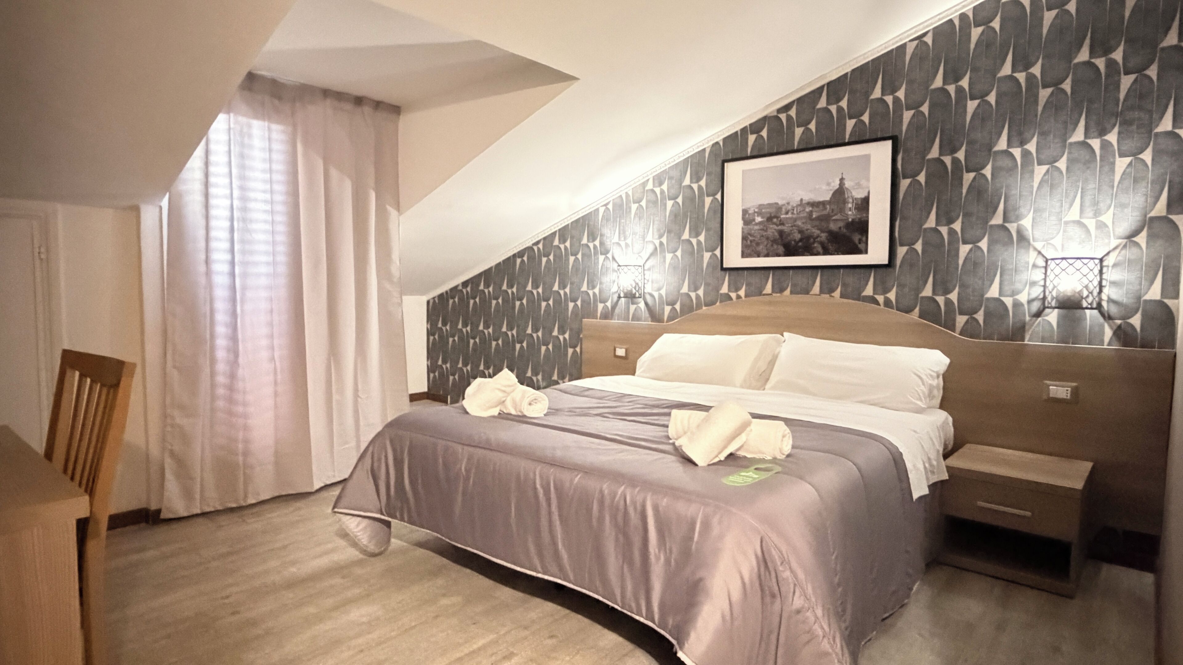 A hotel room with a bed, a nightstand, a chair, a window with curtains, and a wall-mounted picture.