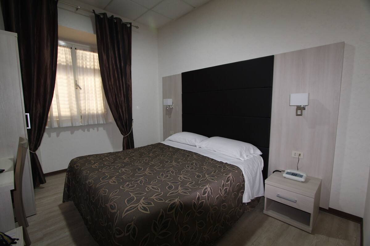 A hotel room with a bed, a nightstand, a lamp, and a window with curtains.