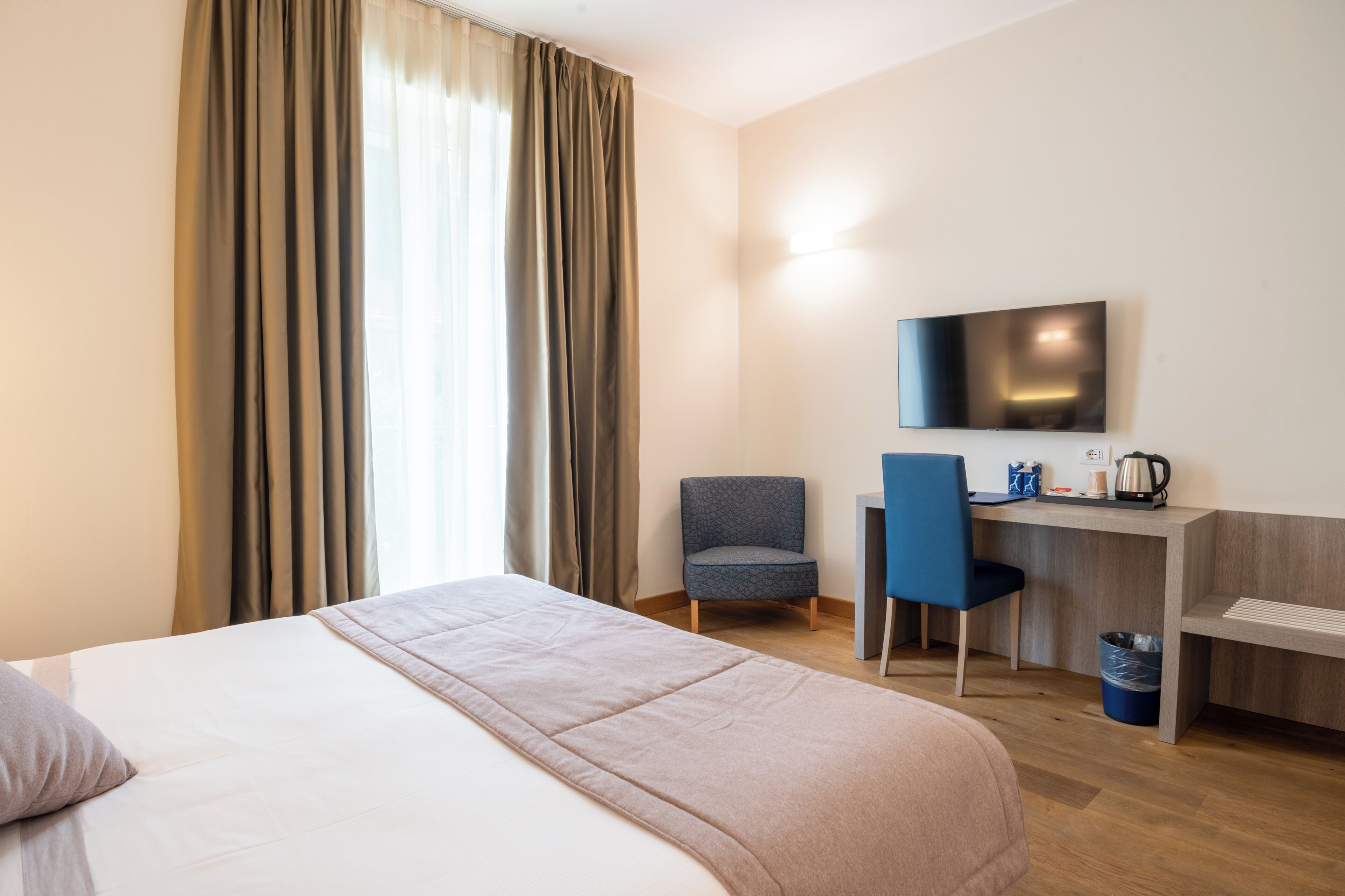Quarto de hotel com cama, escrivaninha, cadeira, televisão e janela com cortinas.