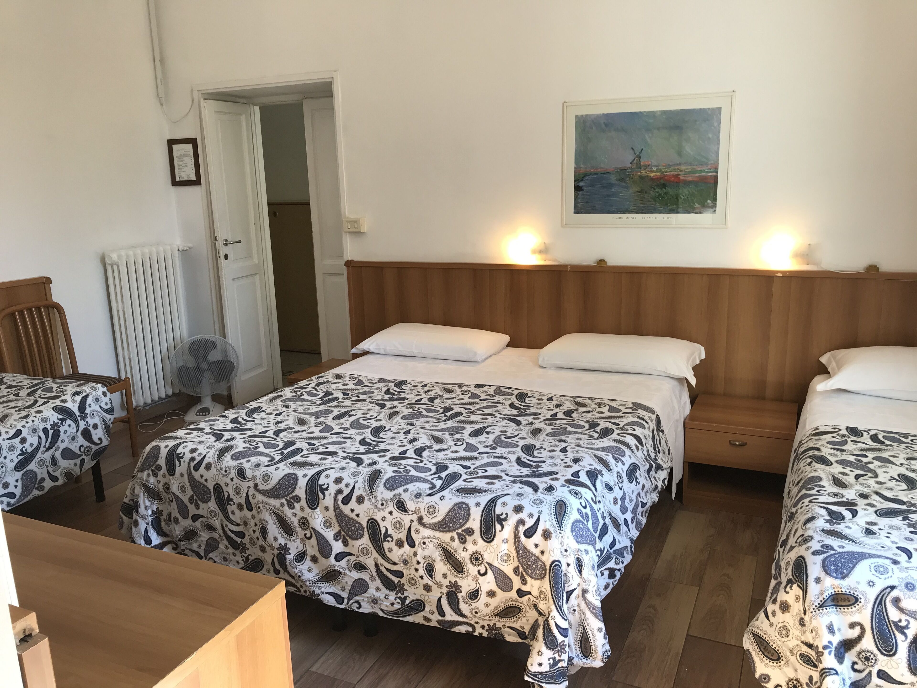 A room with three beds, each with a patterned bedspread, a wooden headboard, and a bedside table. There is a radiator, a fan, and a painting on the wall.
