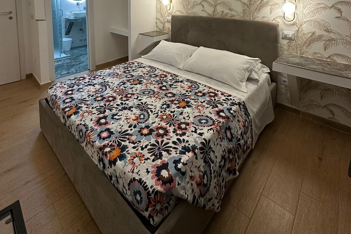 A bedroom with a bed, a nightstand, a wall-mounted light, a patterned wallpaper, and a door leading to a bathroom.