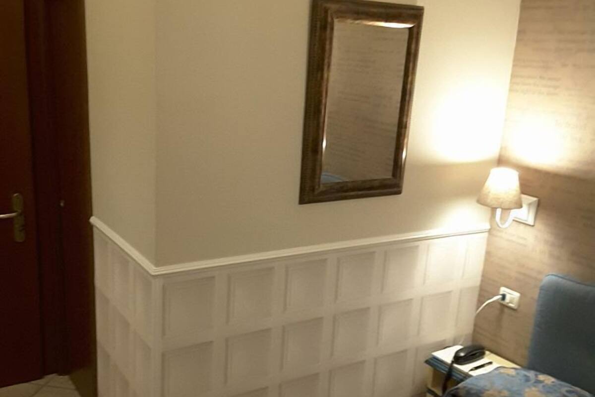 A hotel room with a bed, a nightstand, a lamp, a mirror, and a door.