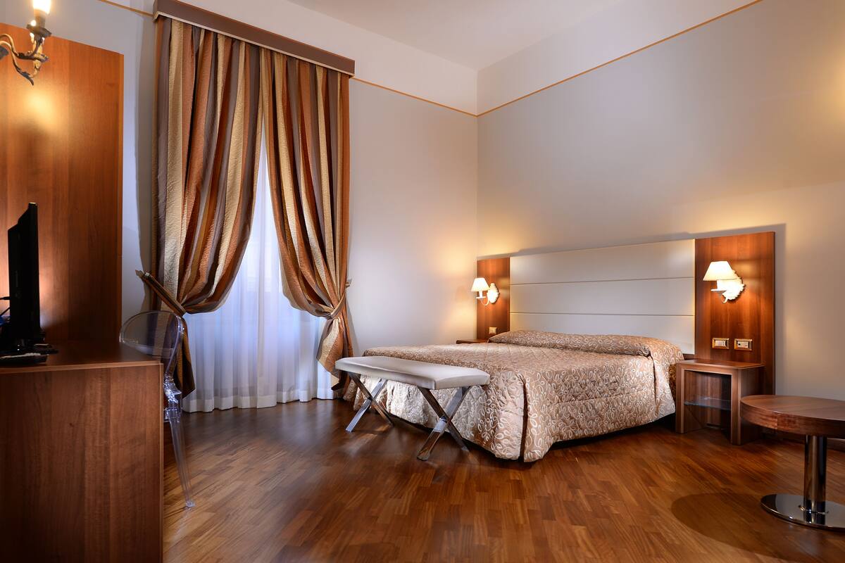 A hotel room with a wooden floor, a bed with patterned bedding, a bench, a TV, and a desk with a lamp.