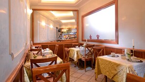 Un restaurante con mesas y sillas de madera, vista a la cocina y un gran ventanal con una vela sobre la mesa.