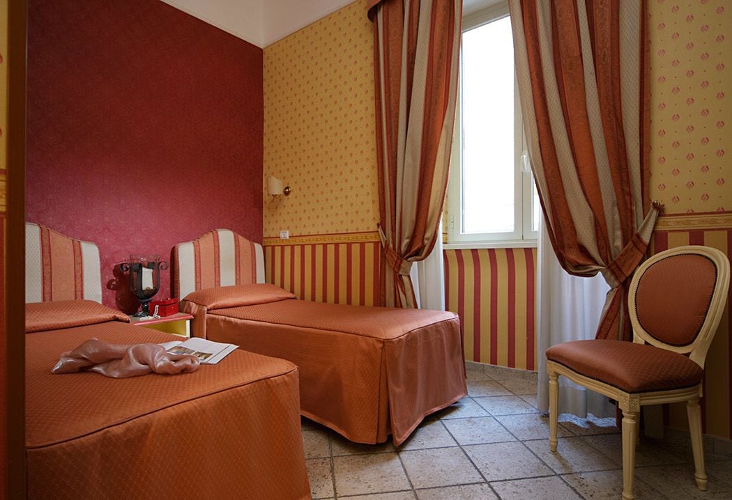 A hotel room with two beds, a chair, a window with curtains, and a patterned wall.