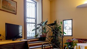 A room with a television on a table, a staircase with a railing, and a window with a view of buildings. - Hotel Monica (Florence)