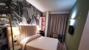 Quarto de hotel com uma cama, uma escrivaninha, uma cadeira, um abajur, um espelho e uma parede com padrão de folhas.