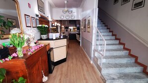 Um saguão de hotel com piso de madeira, um balcão de recepção com um vaso de flores, uma escadaria com degraus de mármore e relógios fixados na parede.