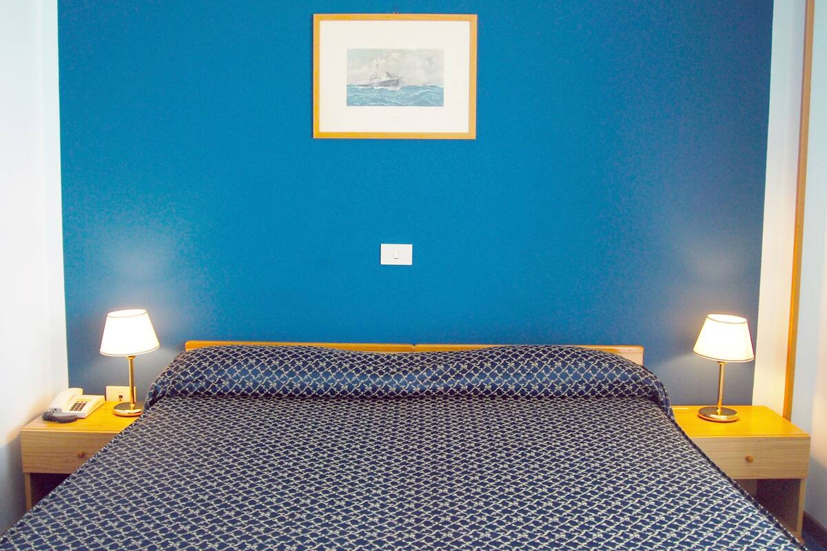 Une chambre d’hôtel avec un mur bleu, un tableau encadré, deux tables de chevet avec des lampes, une couvre-lit à motifs et un téléphone sur la table de gauche.