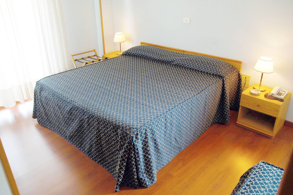 Un lit bien fait, avec une couvre-lit à motifs et des oreillers, une table de chevet avec une lampe et un téléphone, et une chaise en bois.