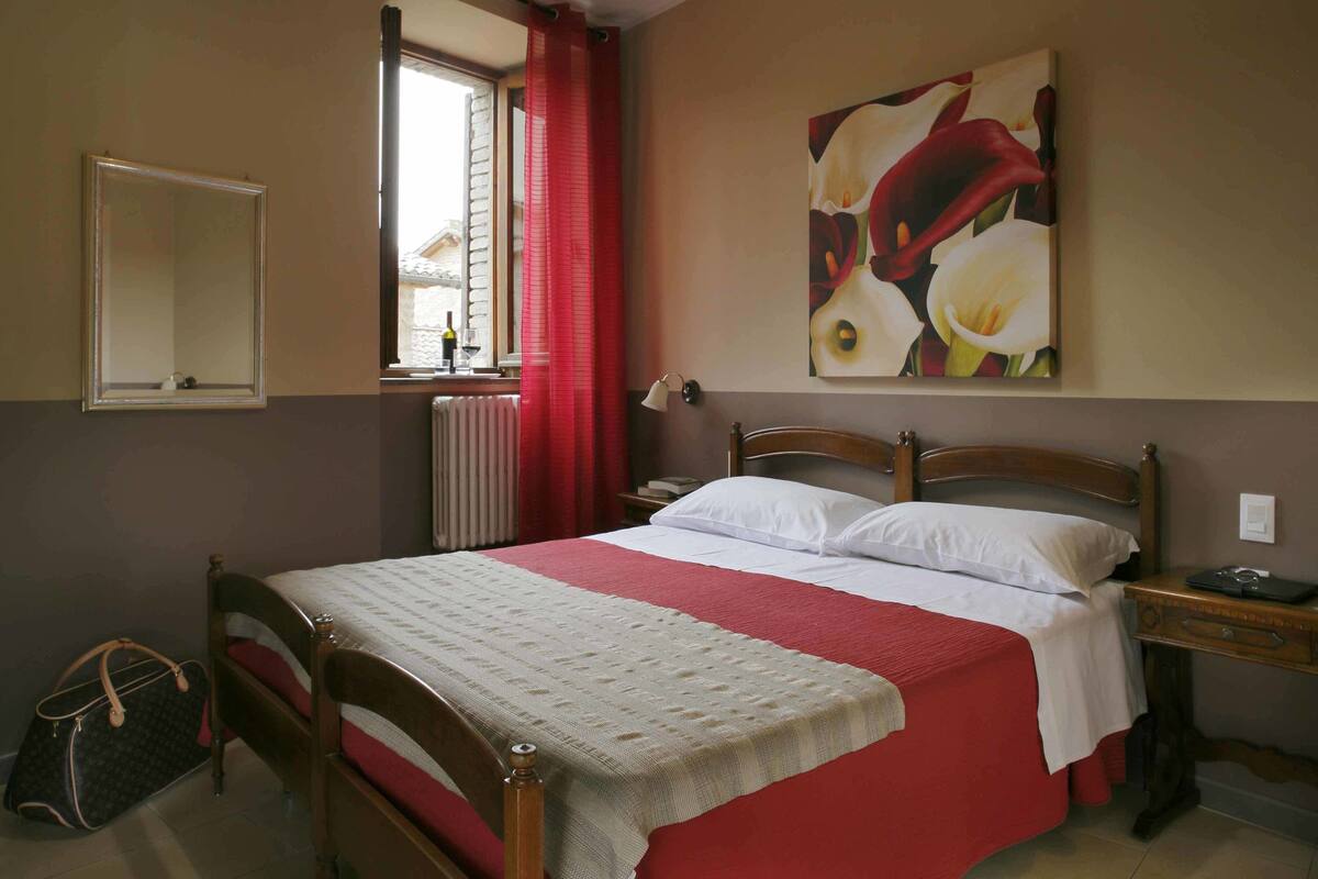 Cama bem arrumada com colcha vermelha e branca, cabeceira de madeira e mesa de cabeceira com abajur.