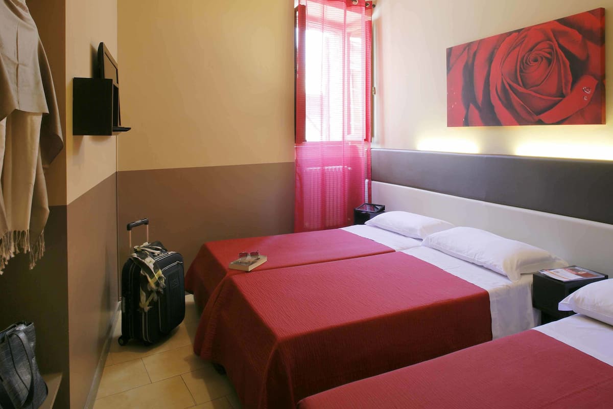 Quarto de hotel com duas camas, um cobreleite vermelho, uma pintura grande de uma rosa, uma mala e uma janela com cortinas cor-de-rosa.