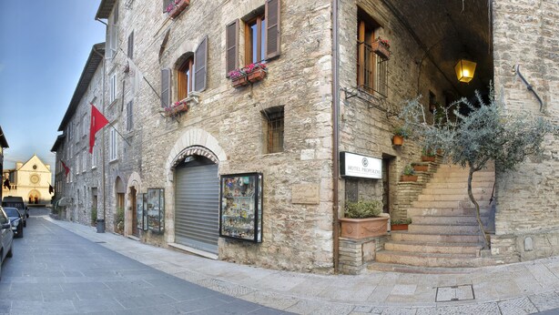 A stone building with a sign for Hotel Properzio, featuring a shop window with souvenirs, a staircase leading to the entrance, and a small tree by the entrance. - Hotel Properzio (Assisi)