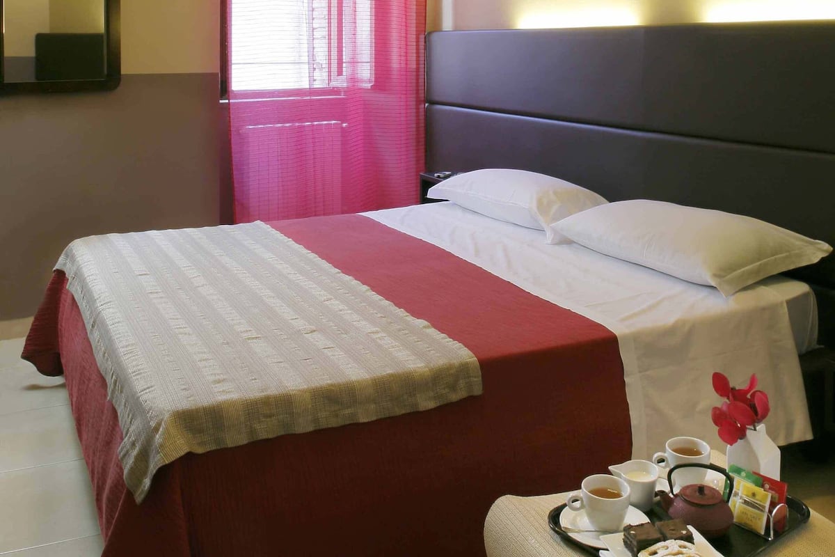 Quarto de hotel com cama, ventoinha no teto, janela com cortinas vermelhas e espelho fixado na parede.