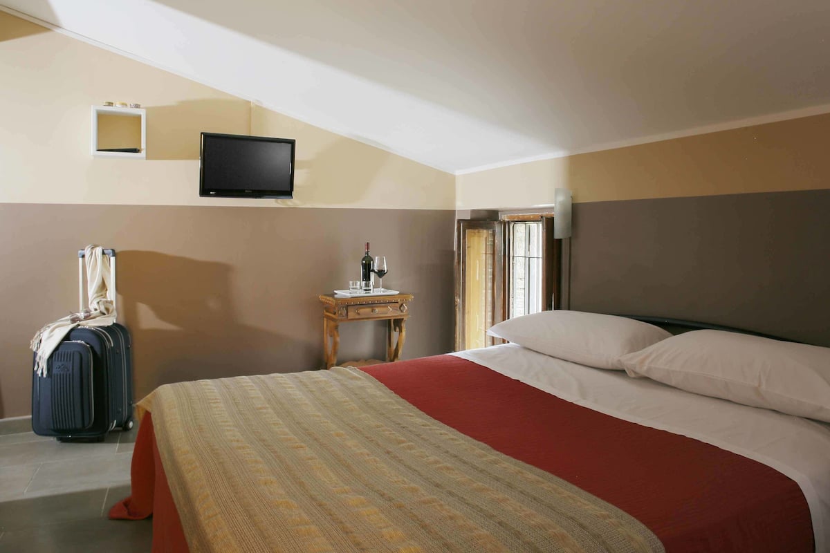 Quarto de hotel com cama, televisão na parede, uma mesinha com garrafa e copos, e uma mala.