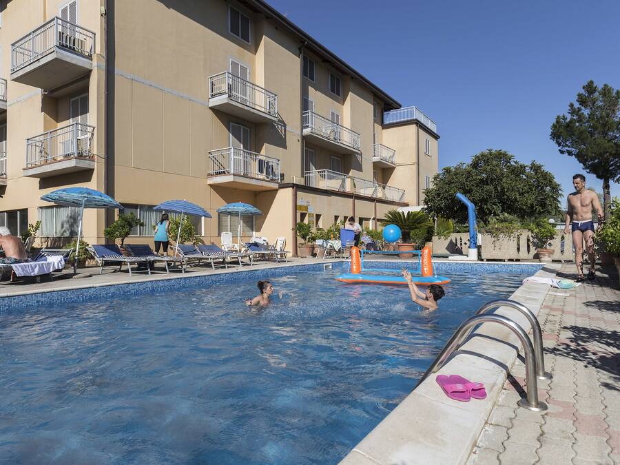 Una piscina con sillas de descanso y sombrillas, rodeada por un edificio de varios pisos con balcones.