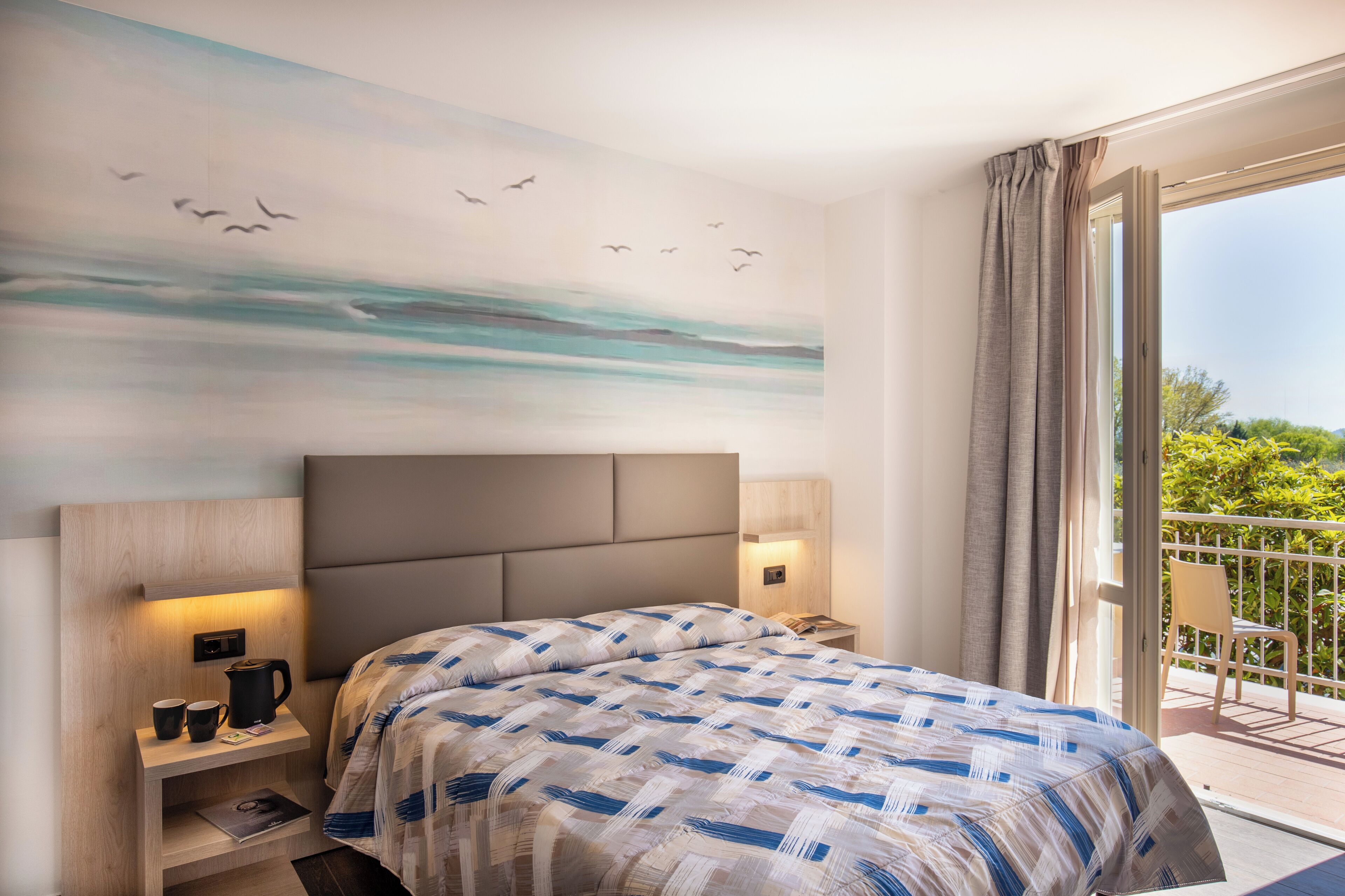 Une chambre d’hôtel comprenant un lit, des tables de chevet, une petite table, une décoration murale et un balcon avec une chaise.