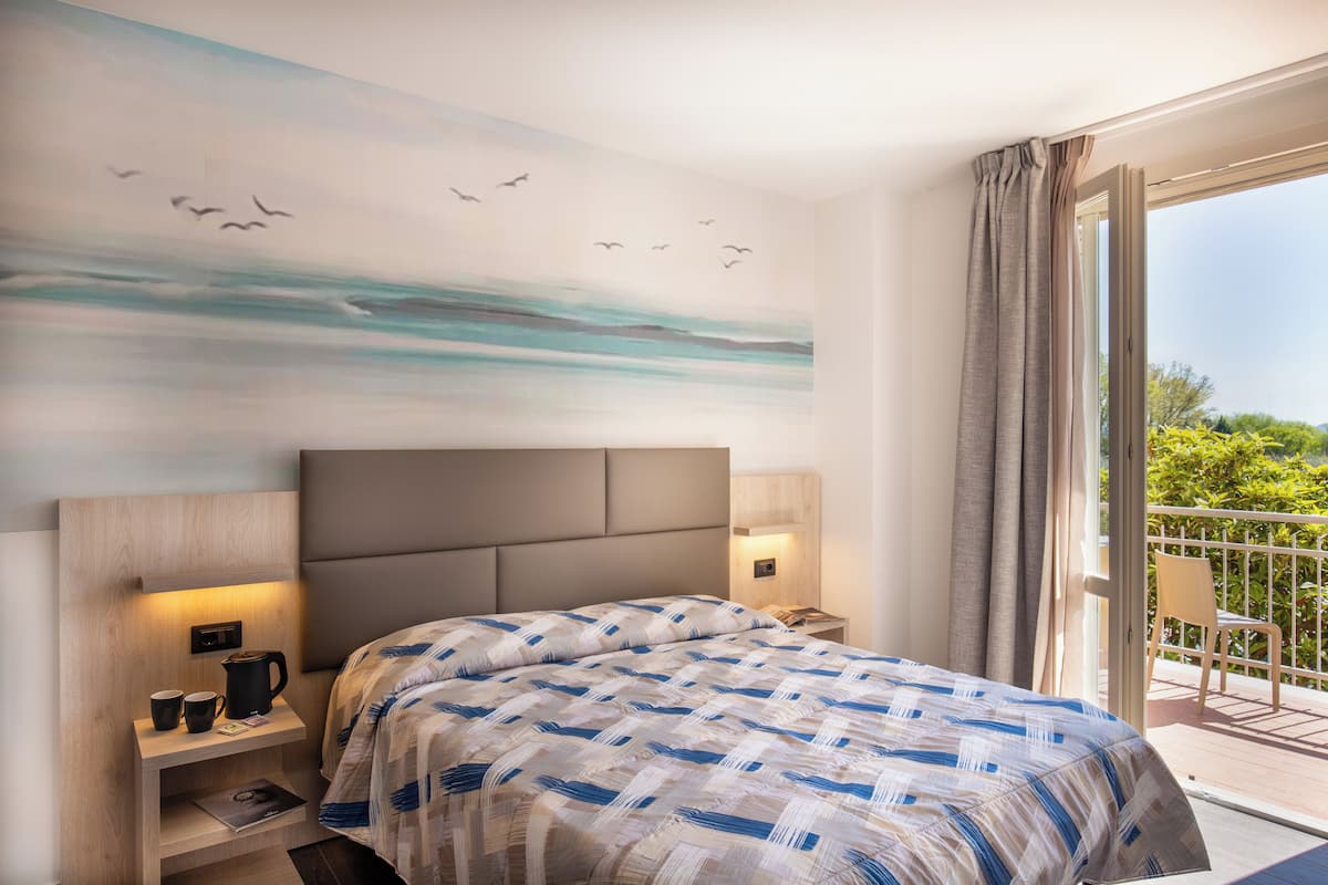 Une chambre d’hôtel comprenant un lit, des tables de chevet, une petite table, une décoration murale et un balcon avec une chaise.
