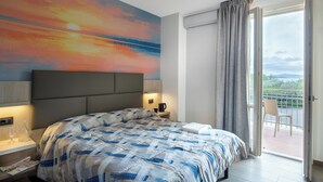 A modern hotel room with a large bed, a nightstand with a lamp, a view of the outdoors through a sliding door, and a painting on the wall.