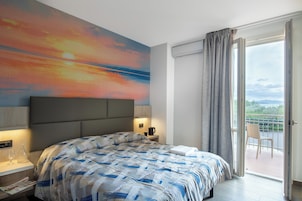 Habitación de hotel moderna con una cama grande, una mesita de noche con lámpara, vista al exterior a través de una puerta corrediza y un cuadro en la pared.