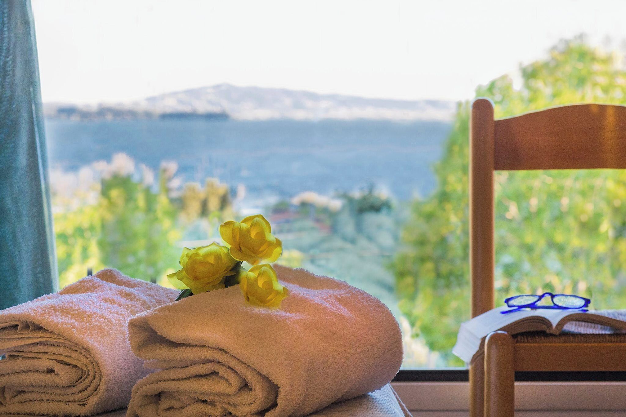 Una ventana con vistas a un paisaje montañoso, una toalla enrollada y un par de gafas sobre una silla de madera.