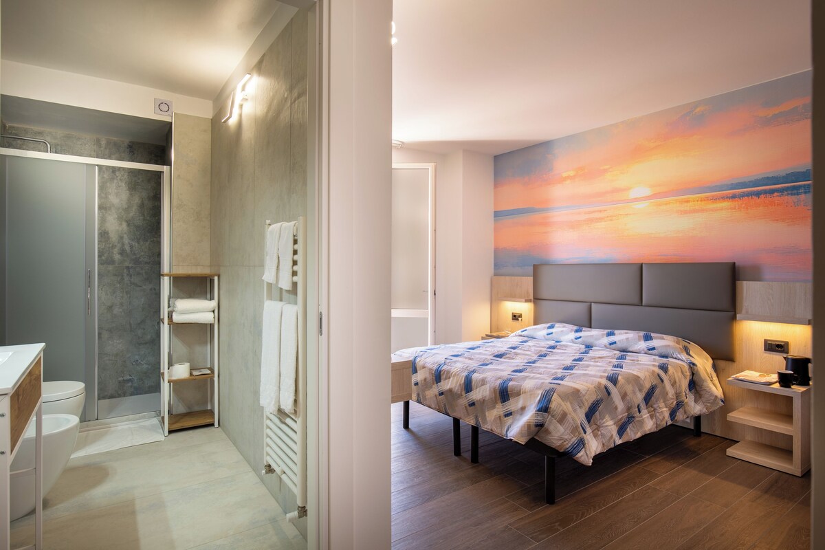 A bedroom with a bed, bedside tables, and a wall with a sunset painting.