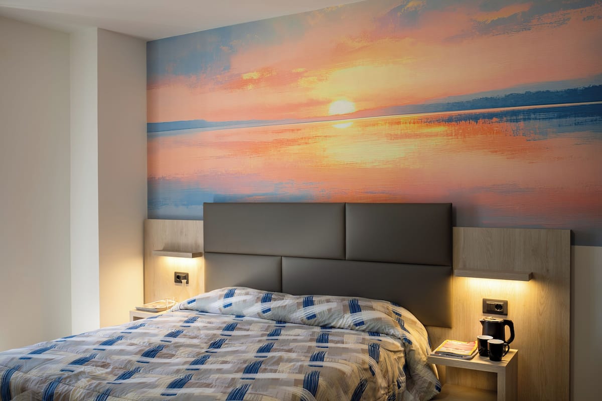 Une chambre d’hôtel avec un lit, des tables de chevet, une petite table et un tableau représentant un coucher de soleil.