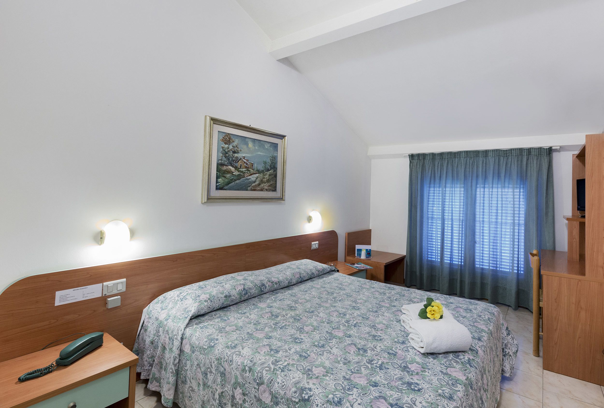Habitación de hotel con cabecera de madera, cama con colcha estampada, mesita de noche con teléfono, lámpara de pared, un cuadro y una ventana con cortinas.