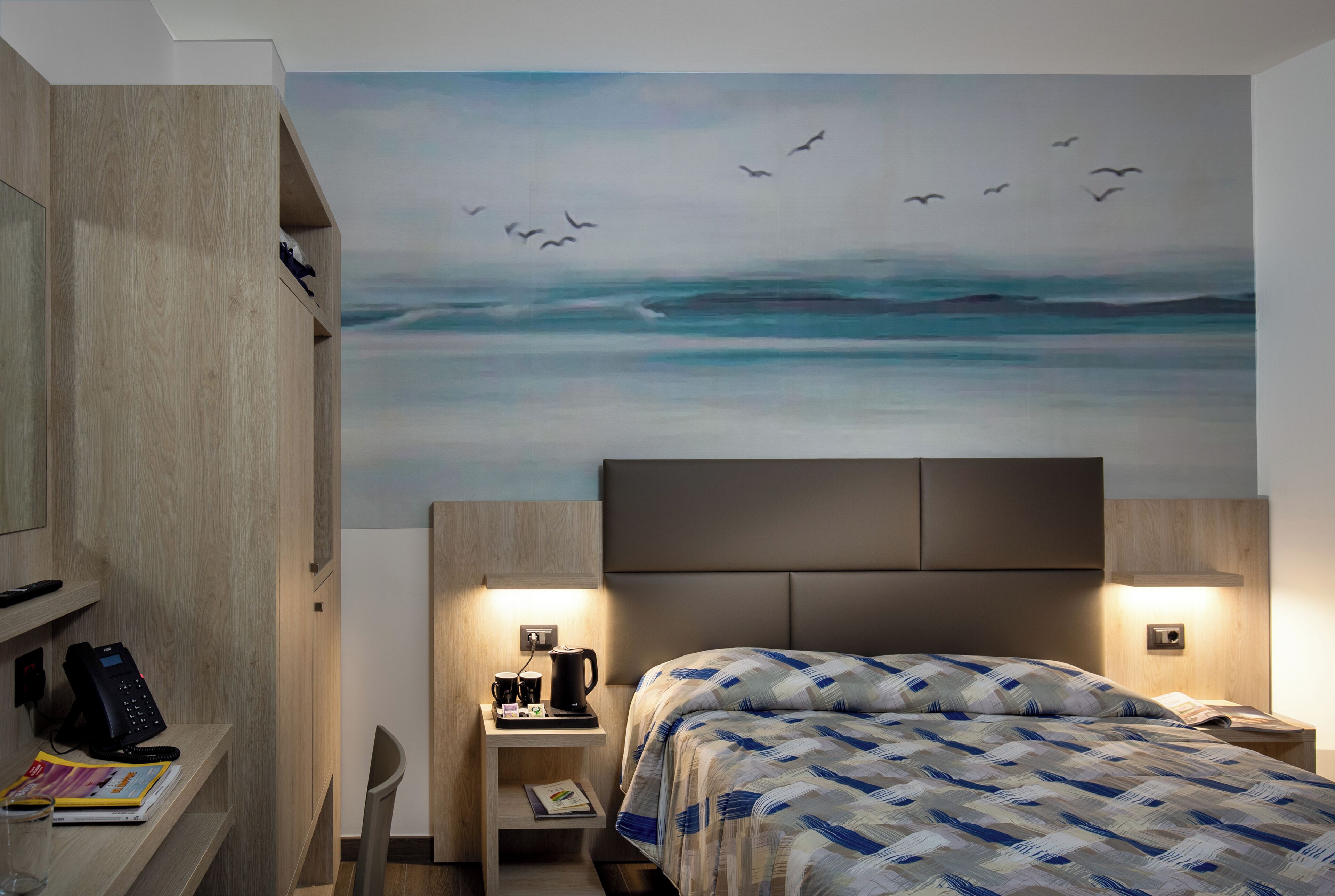 A hotel room with a bed, bedside tables, a desk, a chair, and a wall mural of birds flying over water.
